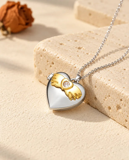 Personalized angel wings heart photo locket necklace in silver tone with gold tone angel wings, resting on a travertine block