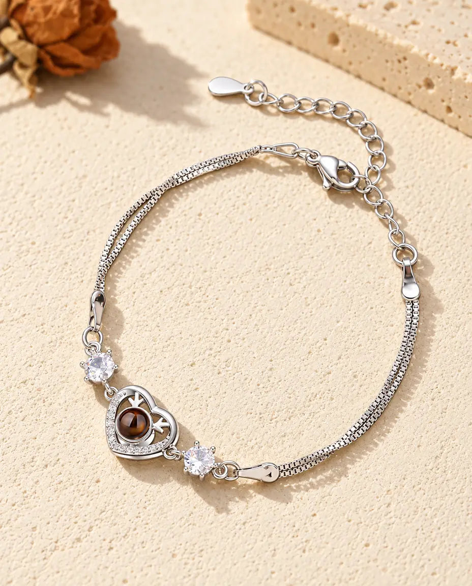 Personalized photo projection bracelet with a silver tone antler heart charm and two cubic zirconia on a double chain, lying on cream plaster