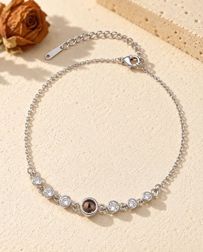 Personalized photo projection bracelet in silver tone with a row of cubic zirconia stones on a fine chain, lying on cream plaster