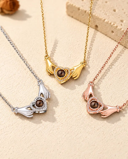 Three colors of the hands holding heart photo projection necklace in a row on cream plaster