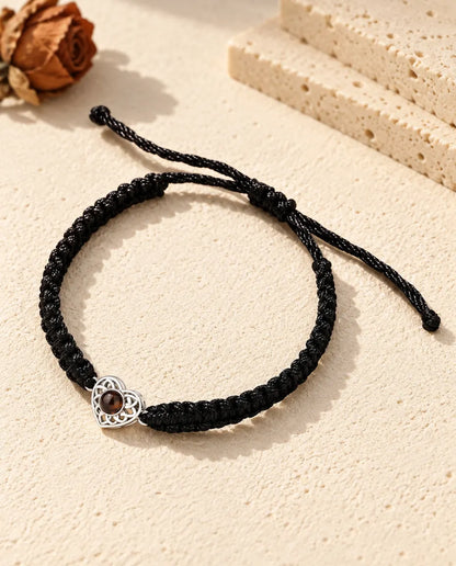 Personalized photo projection bracelet with a silver tone openwork heart charm on a black braided cord, lying on cream plaster