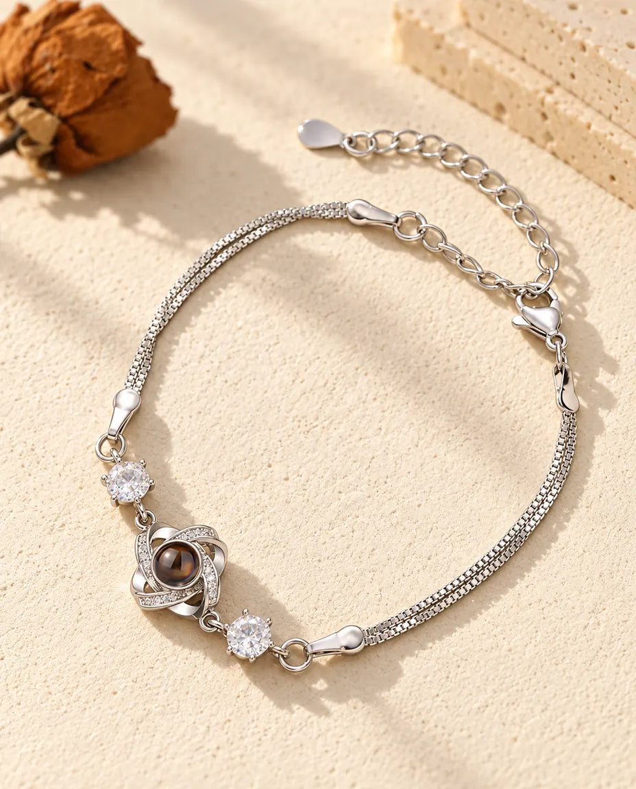 Personalized love knot photo projection bracelet in silver tone with two cubic zirconia on a double box chain, lying on cream plaster