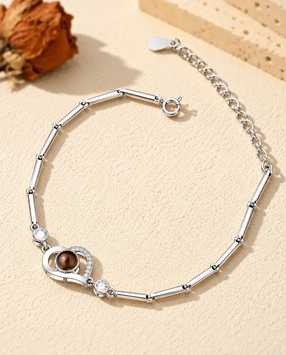 Personalized open heart photo projection bracelet in silver tone on a slim bar chain, lying on cream plaster