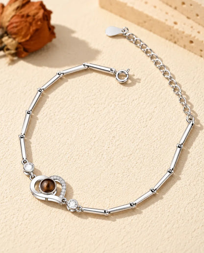Personalized open heart photo projection bracelet in silver tone on a slim bar chain, lying on cream plaster