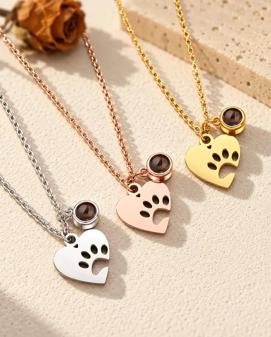Three colors of the paw print heart pet projection necklace on a diagonal on cream plaster