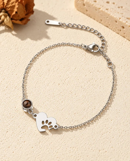 Personalized photo projection bracelet with a silver tone paw print heart tag and projection stone on a fine chain, lying on cream plaster