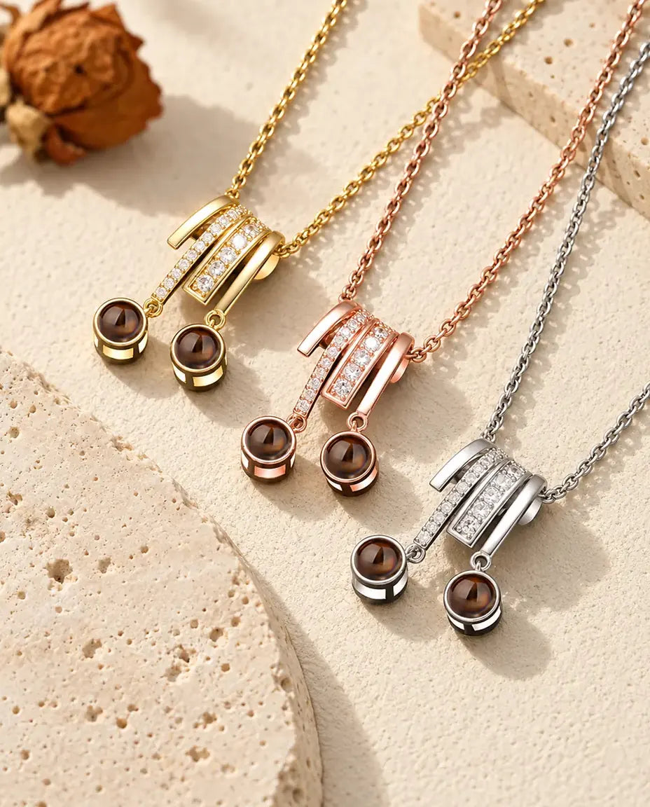 Gold tone, rose gold tone and silver tone two photo projection necklaces on a diagonal beside a travertine slab