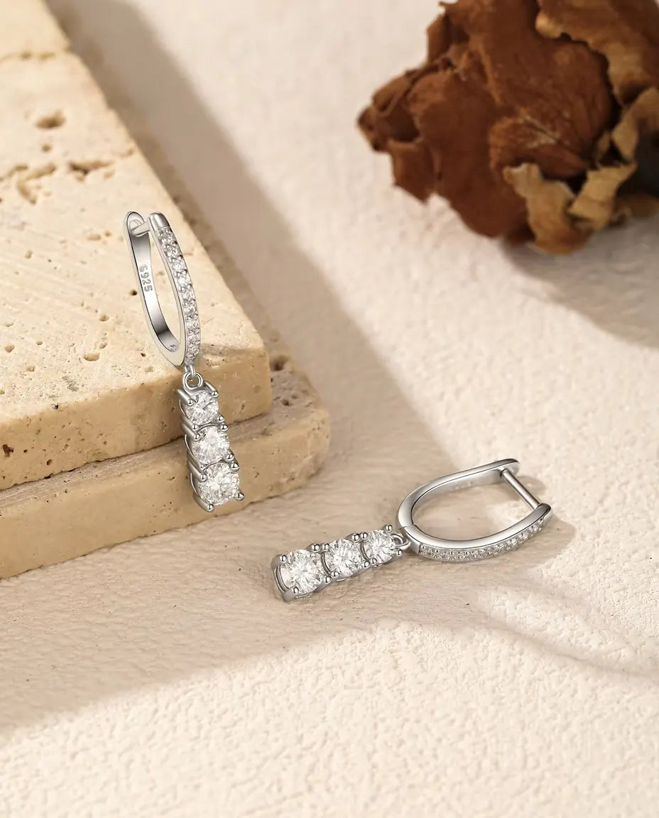 Three Stone Moissanite Drop Hoop Earrings in 925 Sterling Silver - one earring hanging from a travertine slab, the other lying beside it