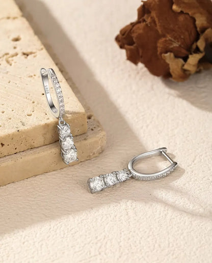 Three Stone Moissanite Drop Hoop Earrings in 925 Sterling Silver - one earring hanging from a travertine slab, the other lying beside it